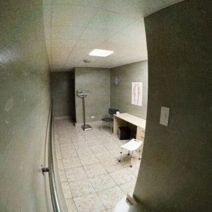 A simple empty medical examination room with desk, chairs, and medical equipment.